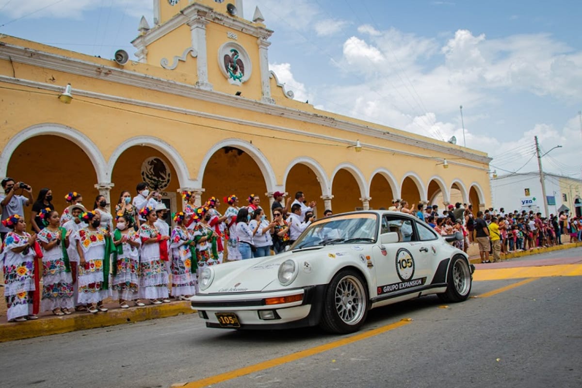 Auto clásico en un rally frente a un palacio municipal, con público en las aceras.